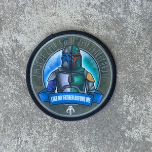 May include: A round, black and green embroidered patch with a Star Wars character, Boba Fett, in the center. The patch has a blue banner with the text "Like my father before me".