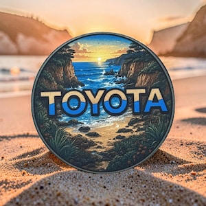 May include: A circular emblem featuring a coastal landscape with cliffs, ocean waves, and a sunset. The word TOYOTA is printed in large, bold, light-colored capital letters across the center, with a blue wave graphic integrated into the text.