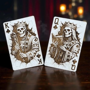 Peut inclure: Deux cartes à jouer représentant des squelettes royaux. Le roi de trèfle tient un sceptre, tandis que la reine de trèfle lit un livre. Les deux cartes ont un aspect vieilli et vintage avec des illustrations marron détaillées sur fond crème.