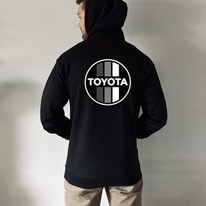 May include: Black hooded sweatshirt with a white and gray Toyota logo on the back. The logo features the word "TOYOTA" in white letters and a circle with four vertical stripes.