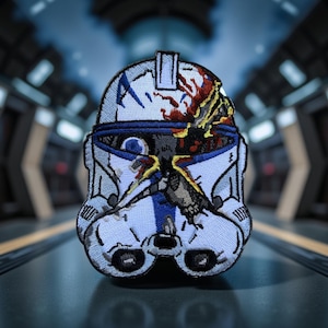 May include: Embroidered patch featuring a white and blue helmet design with battle damage and red accents. The patch has a detailed, stitched appearance, with a blue stripe down the center and black eye details. The background is blurred.