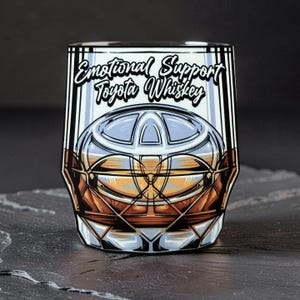 Può includere: Un bicchiere da whisky con un design grafico con il logo Toyota e la scritta "Emotional Support Toyota Whiskey". Il bicchiere ha un motivo geometrico ed è riempito con un liquido marrone.