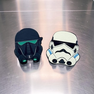May include: Two die-cut Star Wars helmet designs. One is a black helmet with green accents, and the other is a white helmet with black and blue details. The helmets are likely stickers or patches.