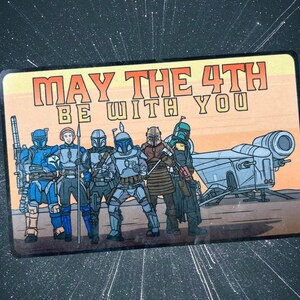 May include: Rectangular graphic featuring a group of characters from a science fiction franchise, with the text "MAY THE 4TH BE WITH YOU." The characters are in various armor and standing in front of a spaceship. The background is a gradient of orange and tan.