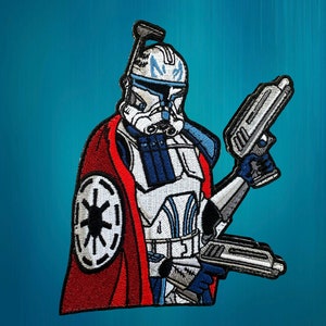 Pode incluir: Um remendo bordado de um clone trooper de Star Wars, vestindo uma capa vermelha e segurando dois blasters. O trooper está usando uma armadura azul e branca com um capacete azul e um símbolo branco no peito.