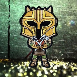 May include: An embroidered patch featuring a cartoon character wearing a helmet and holding two crossed axes, in yellow and brown colours.