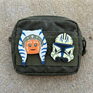 May include: An olive green pouch with two character patches attached. One patch features a cartoon face with blue and white markings, and the other depicts a helmet with yellow and blue accents.