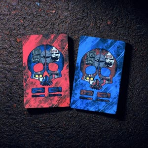 May include: Two rectangular art pieces with a skull design. One is red, the other blue. Each features a skull with a detailed design in the center and two horizontal bars at the bottom. The skull design has a robotic theme.