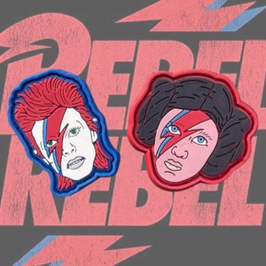 May include: Two red and blue embroidered patches featuring a stylised portrait of David Bowie with a lightning bolt across his face. One patch features a red background and the other a blue background.
