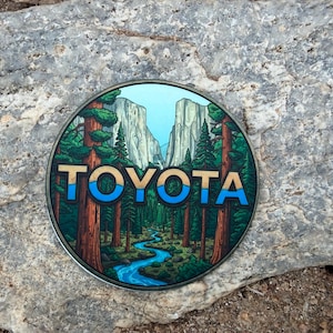 May include: Circular metal emblem with a forest scene, river, and mountains. The word "TOYOTA" is in blue and gold. The emblem is set against a grey stone background. The design evokes a sense of nature and adventure.
