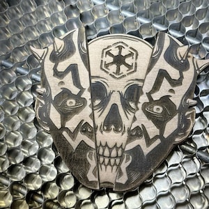 Pode incluir: Um pendurado de parede de madeira preto e branco com um design de caveira e o símbolo do Império de Star Wars. A caveira tem uma expressão ameaçadora com dentes afiados e a testa franzida.