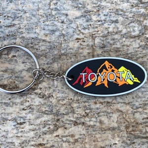 Może przedstawiać: Czarny, owalny brelok do kluczy ze srebrnym kółkiem. Brelok przedstawia stylizowany obraz trzech gór w kolorze czerwonym, pomarańczowym i żółtym z napisem "TOYOTA" białymi literami.