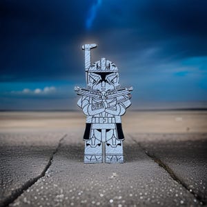 May include: A gray and black wooden figurine of a character from a science fiction franchise. The figurine is in a standing pose, holding two blasters. The background features a cloudy sky and a sandy landscape.