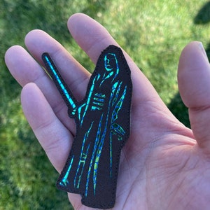 May include: A brown embroidered patch of a figure in a long robe holding a lightsaber. The figure is outlined in blue and green thread.