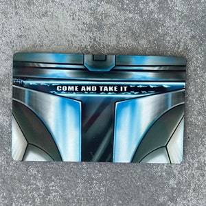 May include: A close-up of a metal card with a blue and silver design. The card has the text "COME AND TAKE IT" printed on it.