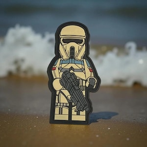 Pode incluir: Um patch bege e preto com um soldado estilizado segurando um rifle blaster. O patch, com uma borda preta, é projetado em um estilo Lego. O fundo mostra uma praia arenosa e ondas.