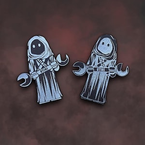 May include: Two silver-colored pins featuring a cartoon character wearing a hooded robe and holding a wrench. The character's name is "UTINI" on the robe.