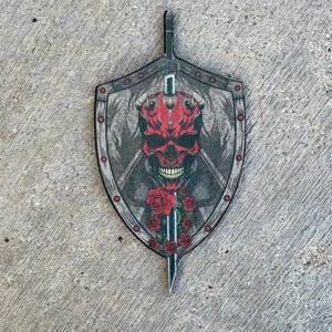 May include: A shield-shaped decorative item featuring a red skull with horns, roses, and a sword design. The shield has a gray border with red accents and a detailed background. The overall design is dark and gothic.