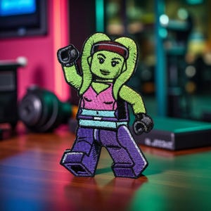 May include: Embroidered patch featuring a green-skinned character with a red headband, pink top, and purple pants. The character is in a dynamic pose, with one arm raised and black gloves. The patch has a cartoonish design and is likely for clothing or accessories.