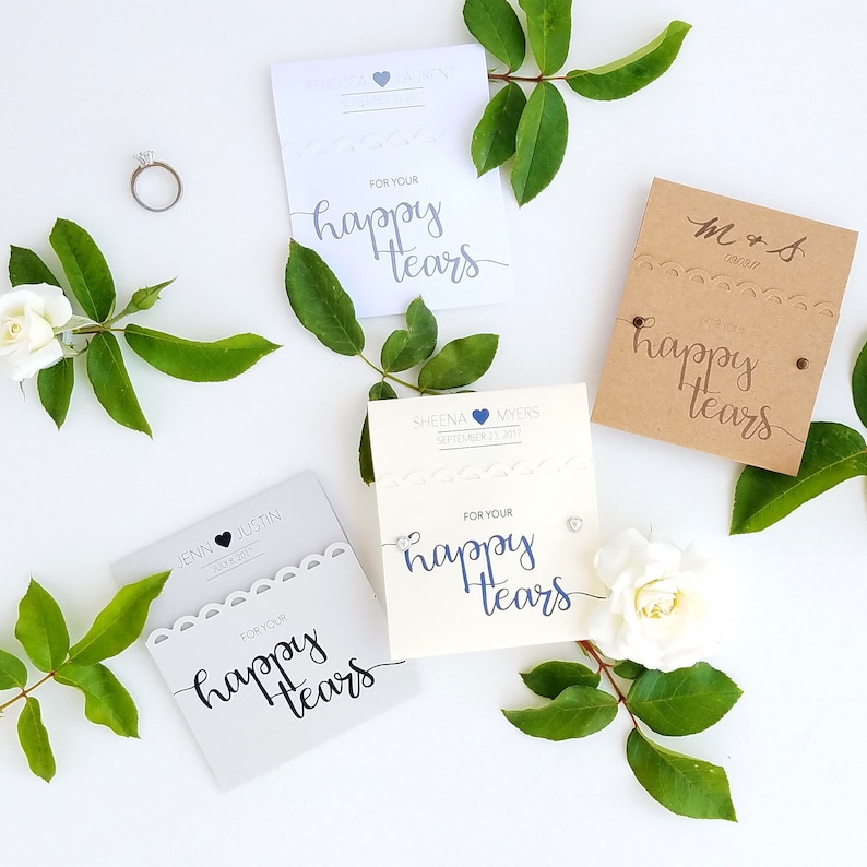 May include: Four white and brown cards with the text "For your happy tears" in black calligraphy. The cards are decorated with lace, hearts, and a silver ring. The cards are surrounded by green leaves and white flowers.