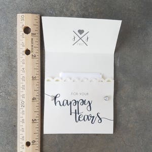 May include: A white card with a scalloped edge and the text "FOR YOUR happy tears" in black script. Two small silver heart-shaped earrings are attached to the card.