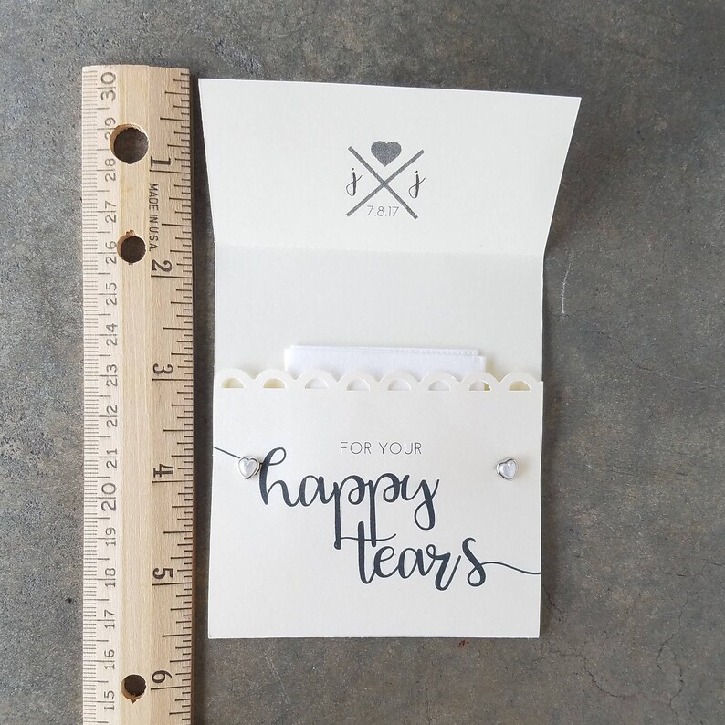 May include: A white card with a scalloped edge and the text "FOR YOUR happy tears" in black script. Two small silver heart-shaped earrings are attached to the card.