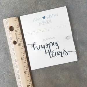 May include: A white card with a decorative border and the text "JENN JUSTIN JULY 8, 2017" and "FOR YOUR happy tears". Two small silver heart-shaped charms are attached to the card.