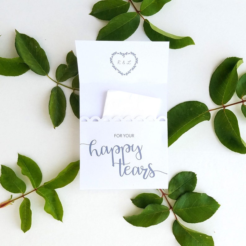 May include: White card with a scalloped edge and the text "For your happy tears" in gray script. A small white tissue is inside the card. The card is surrounded by green leaves.