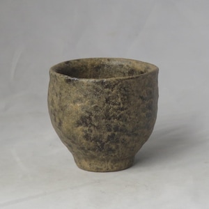 以下が含まれることがあります： 手作りの、質感のあるセラミックカップ。口が広く、底が丸みを帯びています。茶色、タン、グレーのまだら模様の、アースカラーです。表面はざらざらとしていて、自然で有機的な外観です。