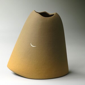 May include: A conical ceramic vase with a matte, golden-brown finish. The vessel features a wide, rounded base that tapers towards an uneven, wavy rim. A small, crescent moon-shaped cutout is positioned on the front surface of the object.