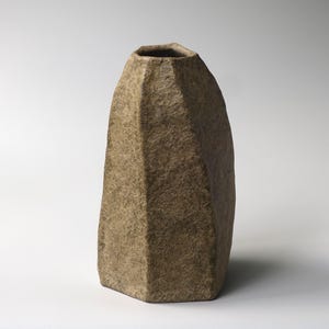 以下が含まれることがあります： 茶色の土のような色合いで、表面がざらざらとした質感の背の高い多面的な花瓶。この容器は、幾何学的で角張ったシルエットが特徴で、上部は狭く開いており、底は平らで、石のようなマットな仕上がりになっています。