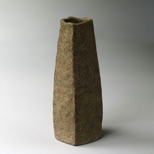 以下が含まれることがあります： くすんだ茶色の土のような色合いで、ざらざらとした質感の表面を持つ、背の高い先細りの花瓶。この容器は、多面的な角張ったボディ、狭い開口部、平らな底面が特徴で、全体的に一貫した風化した外観を見せています。