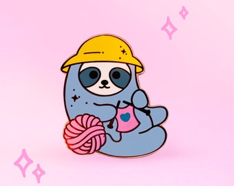 Op de afbeelding: Een blauwe luiaard met een gele helm breit een roze trui met een blauw hart erop. De luiaard houdt roze garen in zijn linkerhand en een breinaald in zijn rechterhand. De luiaard zit op een roze achtergrond.