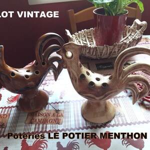 以下が含まれることがあります： 茶色とベージュの釉薬をかけた、ヴィンテージのセラミック製雄鶏の置物2つ。雄鶏は背中に穴が開いており、花瓶として使われていたことを示唆しています。テーブルには「Poteries LE POTIER MENTHON」という文字が見えます。