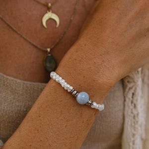 Op de afbeelding: Een delicate armband met kleine witte parels en een lichtblauwe edelsteen kraal. De armband is geaccentueerd met zilverkleurige metalen kralen. Een gouden ketting met een halve maan hanger is ook zichtbaar.