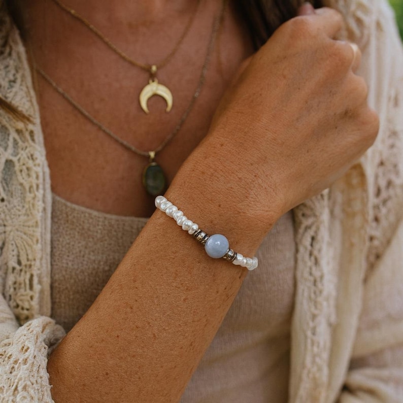 Op de afbeelding: Een delicate armband met kleine witte parels en een lichtblauwe edelsteen kraal. De armband is geaccentueerd met zilverkleurige metalen kralen. Een gouden ketting met een halve maan hanger is ook zichtbaar.