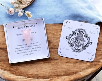 May include: A rose quartz pendulum pendant on a chain inside a white box with printed text about rose quartz, alongside a square white dowsing board featuring a black Hamsa hand illustration and the words Yes, No, and Maybe printed on it.
