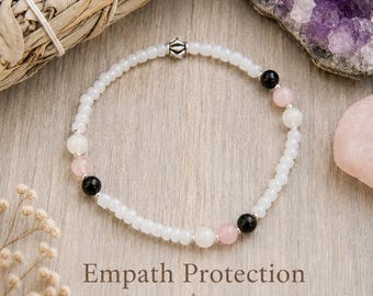 Puede incluir: Una pulsera de cuentas con pequeñas cuentas blancas, intercaladas con cuentas redondas lisas de color negro y rosa pálido, y una cuenta decorativa de metal plateado. El texto Empath Protection está impreso bajo la pulsera en marrón oscuro.