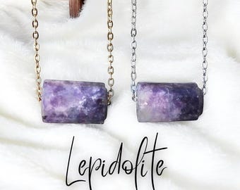 May include: Two necklaces with rectangular lepidolite pendants. One has a gold-colored chain, the other a silver-colored chain. The pendants are a mix of purple and white. The word "Lepidolite" is written in cursive below the necklaces.