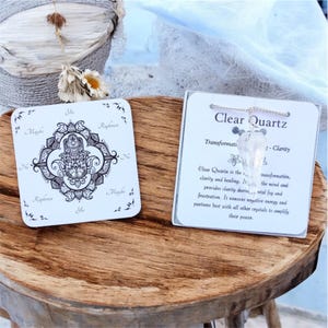 May include: Two white square cards and a clear quartz crystal pendant on a wooden surface. One card features a black and white Hamsa hand design with "Yes", "No", and "Maybe" text. The other card has text that reads "Clear Quartz".