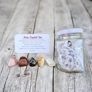 Könnte beinhalten: Ein Aries Kristall-Set. Ein klares Glas mit weißem Deckel ist mit weißem Papier gefüllt. Fünf polierte Steine in Rosa, Braun, Schwarz, Gelb und Weiß werden neben einer Karte mit Text angezeigt. Die Steine sind für den Widder.