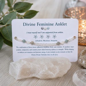 May include: An anklet featuring small, round, multi-colored beads including shades of green, white, and grey, displayed on a white card labeled Divine Feminine Anklet. The card includes text about Labradorite, Moonstone, and Amazonite stones.