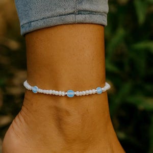Pode incluir: Tornozeleira de contas brancas com três contas azuis claras. A tornozeleira é usada em um tornozelo, com um fundo desfocado de folhagem verde e uma pessoa usando jeans azuis.