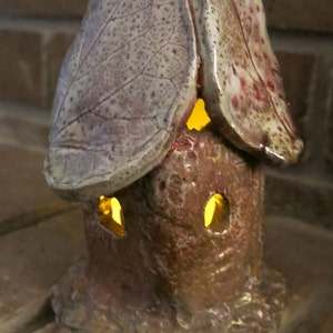 May include: A small ceramic house with a roof made of two leaves. The house has a light inside, illuminating the windows. The house is painted in shades of brown, white, and red.