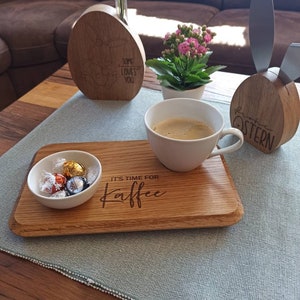 Könnte beinhalten: Ein Holztablett mit der Gravur "It's time for Kaffee". Auf dem Tablett stehen eine Tasse Kaffee, eine kleine Schale mit Pralinen und eine Holzhasenfigur mit der Aufschrift "Frohe Ostern".