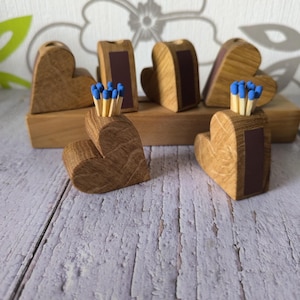 Könnte beinhalten: Herzförmige Streichholzhalter aus Holz mit blauköpfigen Streichhölzern. Die Halter sind aus hellem Holz gefertigt und haben einen dunkelbraunen rechteckigen Bereich. Mehrere Halter sind auf einer Holzoberfläche angeordnet, was auf einen dekorativen oder funktionalen Artikel hindeutet.