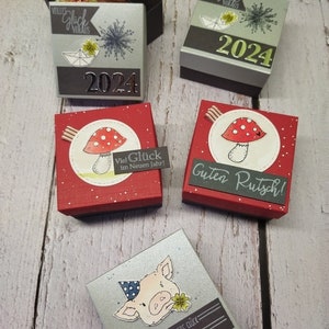 Könnte beinhalten: Vier kleine Geschenkboxen mit verschiedenen Designs. Die erste Box ist silber mit einer goldenen 2024 und dem Text "volles Glück voraus". Die zweite Box ist rot mit weißen Punkten und einem Pilz mit dem Text "Viel Glück im Neuen Jahr!" Die dritte Box ist rot mit weißen Punkten und einem Pilz mit dem Text "Guten Rutsch!" Die vierte Box ist silber mit einem Schwein, das einen Partyhut trägt und dem Text "Gesundheit Liebe Glück Frohes Neues Jahr".