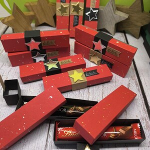 Könnte beinhalten: Rote und schwarze Geschenkboxen mit goldenen Sternen und dem Text "glückliche Weihnachtsgrüße" auf einer weißen Holzoberfläche. Die Boxen sind mit Schokoladentafeln gefüllt.