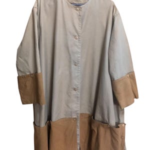 May include: A long, button-down coat in a neutral color palette. The coat features tan fabric with brown accents on the cuffs and pockets. The coat is hanging on a black hanger.