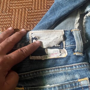 May include: Close-up of blue denim jeans with a worn appearance. The back pocket area is visible, with a label showing size measurements and the text "Mission No. 503". The jeans have a classic five-pocket design and yellow stitching.